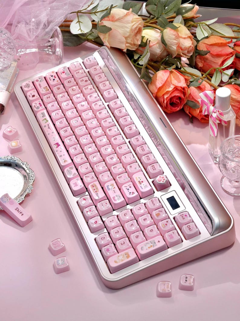 May include: A pink mechanical keyboard with cat-themed keycaps and a silver frame. The keyboard has a small digital display. Additional keycaps are scattered on a pink surface. The text "CAT BUTTERFLY" is visible.