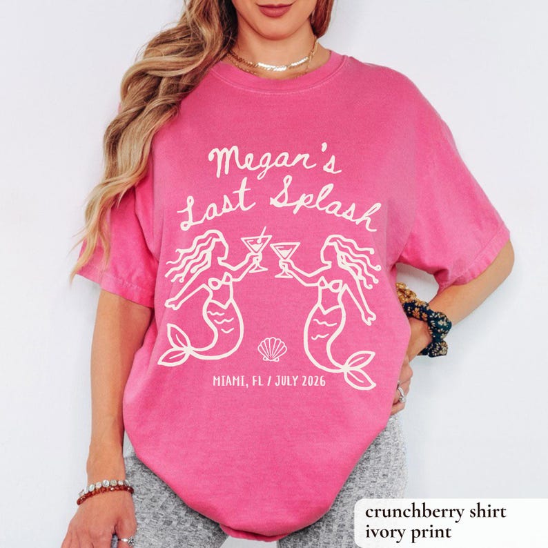 May include: A crunchberry pink t-shirt with the text "Megan's Last Splash" in white script. The design includes two mermaid illustrations toasting cocktails. The text "Miami, FL / July 2026" is printed below the mermaids.