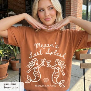 May include: A yam-colored t-shirt with an ivory print. The shirt features the text "Megan's Last Splash" above two mermaid illustrations toasting cocktails. The text "Miami, FL / July 2026" is printed below the mermaids.