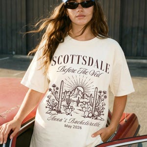May include: Cream-colored t-shirt with a desert landscape graphic. The design includes cacti, mountains, and a rising sun, with the text "SCOTTSDALE Before The Veil" and "Anna's Bachelorette May 2026".