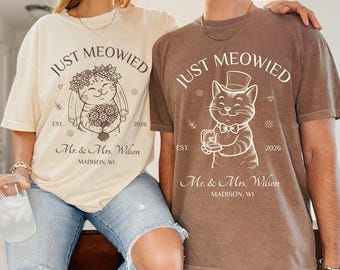 May include: Two matching beige and brown t-shirts with the text "JUST MEOWIED" and a cat illustration. The beige shirt has a cat wearing a floral crown and holding flowers. The brown shirt has a cat wearing a top hat and holding a ring.