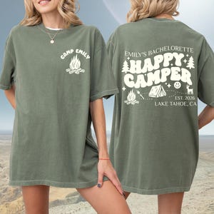 Peut inclure: T-shirt vert olive avec texte et graphiques blancs. Le devant affiche "Camp Emily" avec un motif de feu de camp. Le dos indique "Emily's Bachelorette Happy Camper" avec des graphiques de camping et "Est. 2026 Lake Tahoe, CA."