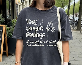 May include: Dark gray t-shirt with white text that reads "They Caught Feelings." Below the text is "I caught this t-shirt, Chris and Danielle 10.04.2026." The shirt also features two champagne glasses. The shirt is a casual style.