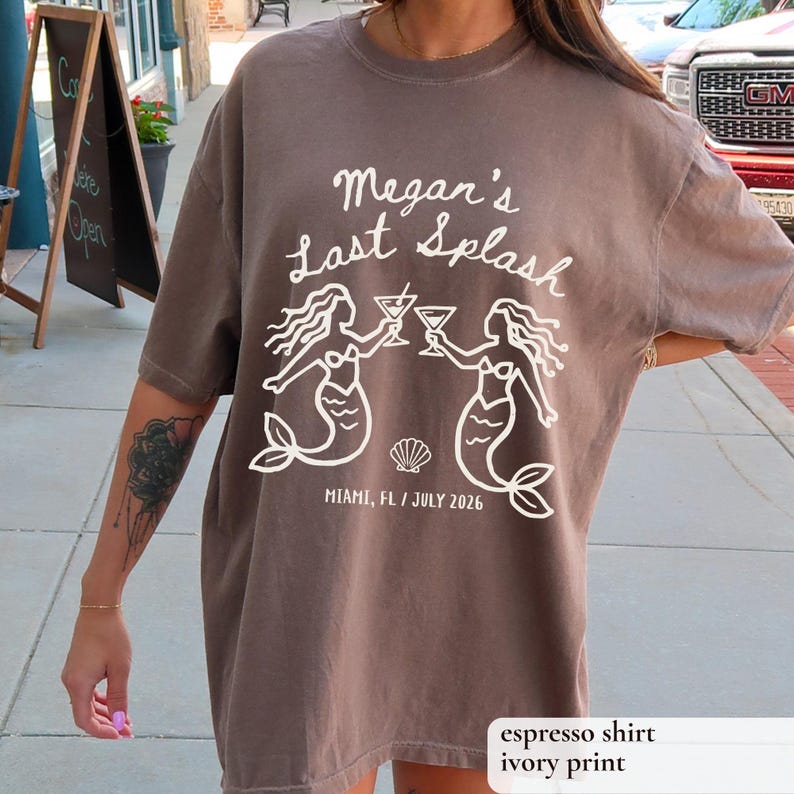 May include: Espresso-colored t-shirt with white text and graphic. The text reads "Megan's Last Splash" above two mermaid illustrations holding cocktails. Below, it says "Miami, FL / July 2026".