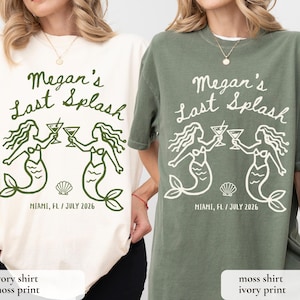 May include: Ivory and moss green t-shirts with a graphic design. The design features the text "Megan's Last Splash" above two mermaid illustrations holding cocktails. Below the mermaids is the text "MIAMI, FL / JULY 2026".
