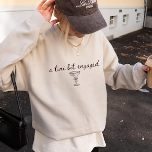 May include: Beige sweatshirt with the text "a tini bit engaged" and a martini glass illustration. The sweatshirt is paired with a dark brown baseball cap with white text and gold necklaces. A black purse hangs on the left side.