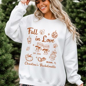May include: A white long-sleeve sweatshirt featuring brown graphic illustrations of a coffee cup, disco ball, champagne glasses, a pumpkin, and rings. The text reads Fall in Love, EST. 2026, MIAMI, FL, and Christina's Bachelorette in a script font.