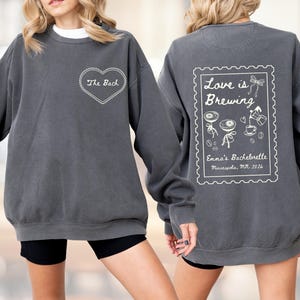 May include: Charcoal gray sweatshirt with white text and graphics. The front features a heart with "The Bach" inside. The back has a stamp-like design with "Love is Brewing" and "Emma's Bachelorette, Minneapolis, MN 2026".