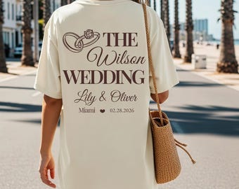 May include: Cream-colored t-shirt with a brown graphic of two intertwined rings and the text "THE Wilson WEDDING" above "Lily & Oliver" and the date 02.28.2026. The word "Miami" is below the names. The shirt is worn by a person.