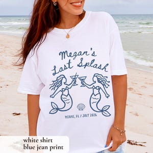 May include: White t-shirt with a blue graphic that reads "Megan's Last Splash" with two mermaid illustrations toasting cocktails. The shirt also includes the text "Miami, FL / July 2026".