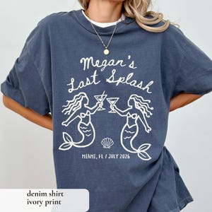 May include: Denim blue t-shirt with the text "Megan's Last Splash" in a white script font. The shirt features a graphic of two mermaids toasting cocktails, with the location and date below. The shirt is a relaxed fit.