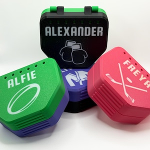 May include: A selection of personalised sports mouthguard cases in green, black, purple, and pink. Each case displays a name and a sport graphic: rugby ball, boxing gloves, judo uniform, and hockey sticks. The cases are designed to hold mouthguards.