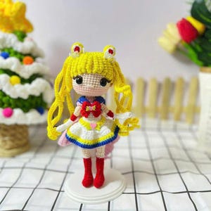 Puede incluir: Muñeca de ganchillo hecha a mano con un personaje de pelo amarillo con un atuendo de marinero. La muñeca tiene un vestido blanco con ribetes azules y amarillos, botas rojas y está sobre un soporte blanco. La muñeca mide aproximadamente 15 cm de alto.