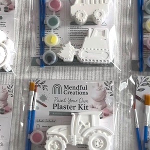 Könnte beinhalten: Ein Mendful Creations Gips-Malset in transparenter Kunststoffverpackung. Das Set enthält zwei weiße Gipsfiguren von Baufahrzeugen oder Traktoren, zwei Pinsel mit blauem Griff und vier kleine, runde Farbtöpfe in verschiedenen Farben.