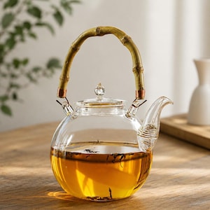 Puede incluir: Una tetera de vidrio transparente con un asa de bambú y una tapa de vidrio. La tetera está llena de té de color ámbar. El asa está hecha de un trozo de bambú curvado. El pico tiene un diseño curvo.