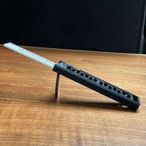 May include: A black and white plastic tool with a series of small teeth along one edge. The tool is angled, resting on a wooden surface. A small, brown cylindrical object supports the tool.