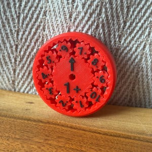 May include: A red, circular 3D-printed gear-based clock with black numerals and an arrow. The clock has a series of interlocking gears around the perimeter and the numbers 1 through 10. The clock is on a wooden surface.