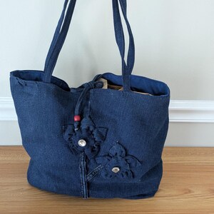 May include: A dark blue denim tote bag featuring two fabric flower appliqués on the front, each centered with a clear rhinestone. The bag includes two long shoulder straps, a drawstring closure with a single red bead, and a dark blue fabric lining.
