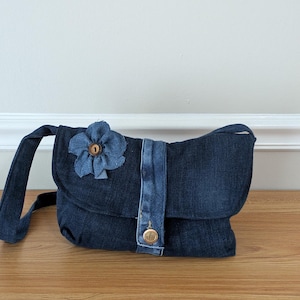 May include: A dark blue denim shoulder bag featuring a fold-over flap with a vertical denim strap and a metallic button closure. The bag is decorated with a fabric flower accent made from frayed denim material with a small brown button in the center.