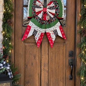 May include: A large fabric bow decoration featuring a patchwork design with red, green, white, and black colors. The fabric patterns include snowflakes, plaid, and foliage motifs. The bow has two large top loops and two long, pointed tails hanging down.