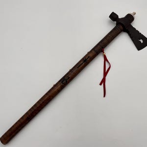 May include: A handcrafted tomahawk with a dark wooden handle and a black axe head. The handle features carved designs and a red tassel. The tomahawk is a traditional Native American tool and weapon.