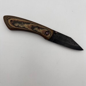 May include: A folding knife with a textured, brown and tan handle and a black, patterned blade. The blade has a unique, angular shape. The handle is secured with three rivets. The knife is shown against a white background.