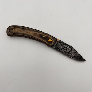 May include: A folding knife with a patterned blade and a wooden handle. The handle is a light brown color with darker brown marbling. The blade has a dark, swirling pattern. Small gold-colored rivets secure the handle.