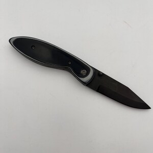May include: A black folding knife with a black blade and a black and gray handle. The knife is open and angled, showcasing its sharp edge. The handle has a textured appearance.