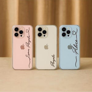 以下が含まれることがあります： パステルカラーのピンク、クリーム、ブルーの3つのパーソナライズされた携帯電話ケース。各ケースは透明なボーダーで、エレガントなスクリプトで名前が書かれています。ピンクのケースには「Lana Angela」、クリームのケースには「Angela」、ブルーのケースには「Alice」と書かれています。
