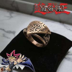 Puede incluir: Un anillo de metal en tono bronce con el símbolo del Ojo de Anubis en su cara frontal y grabados intrincados a lo largo de la banda. El logotipo de Yu-Gi-Oh! es visible en la esquina superior derecha y una ilustración de personaje abajo.
