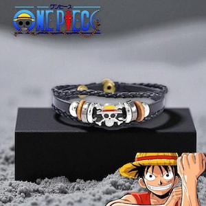 以下が含まれることがあります： 黒い革のブレスレット。シルバーのドクロとクロスボーンのチャーム、黄色と赤の帽子、木と金属のアクセントが特徴です。ブレスレットは、背景に「ONE PIECE」のロゴが入った黒い箱に展示されています。