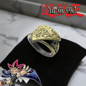 Pode incluir: Um anel dourado com um design de olho, sobre um fundo preto. O anel apresenta detalhes intrincados e provavelmente é inspirado na série Yu-Gi-Oh!. Uma peça de joalharia única.