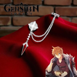 Puede incluir: Broche plateado con temática de Genshin Impact. El broche presenta un diseño floral, una gema roja y una cadena. El logotipo de Genshin Impact es visible en el fondo. El broche se muestra sobre una superficie de terciopelo rojo.