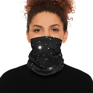 Op de afbeelding: Een zwarte nekwarmer met een sterrenhemelontwerp met heldere witte sterren. De nekwarmer wordt om de nek gedragen en bedekt de onderkant van het gezicht. De persoon op de foto draagt een zwart shirt.