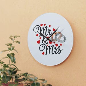 Peut inclure: Une horloge ronde blanche avec l'inscription "Mr & Mrs" en noir, entourée de cœurs rouges. Deux alliances sont représentées près des aiguilles. L'horloge est accrochée à un mur orange clair.