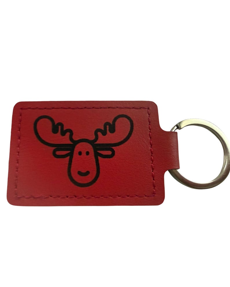 May include: Red leather keyring featuring a black cartoon moose design. The keyring has a rectangular shape with a stitched border and a silver metal ring attached.