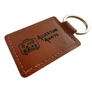 May include: A brown leather keychain with a silver key ring. The rectangular tag features the words "Adventure Awaits" and a graphic of a suitcase, trees, and a tent. The edges are stitched with a lighter brown thread.