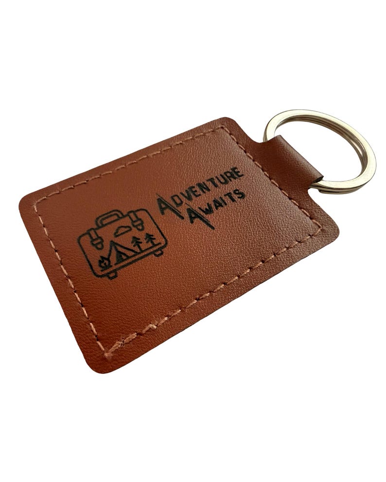 May include: A brown leather keychain with a silver key ring. The rectangular tag features the words "Adventure Awaits" and a graphic of a suitcase, trees, and a tent. The edges are stitched with a lighter brown thread.