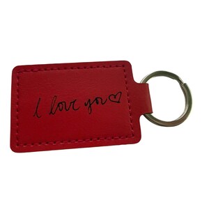 May include: A red rectangular keyring with the handwritten phrase "I love you" in black script, with a heart symbol. It features a silver metal ring for attaching keys.