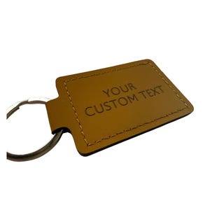 May include: A tan leather keyring with the words "YOUR CUSTOM TEXT" embossed on the front. It has a silver metal ring for attaching keys. The edges are stitched with a contrasting thread.