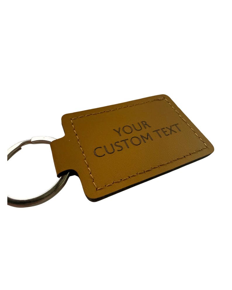 May include: A tan leather keyring with the words "YOUR CUSTOM TEXT" embossed on the front. It has a silver metal ring for attaching keys. The edges are stitched with a contrasting thread.
