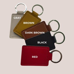 May include: A selection of rectangular keyrings in shades of grey, brown, dark brown, black, and red. Each keyring has a metal ring and the colour name printed on it.