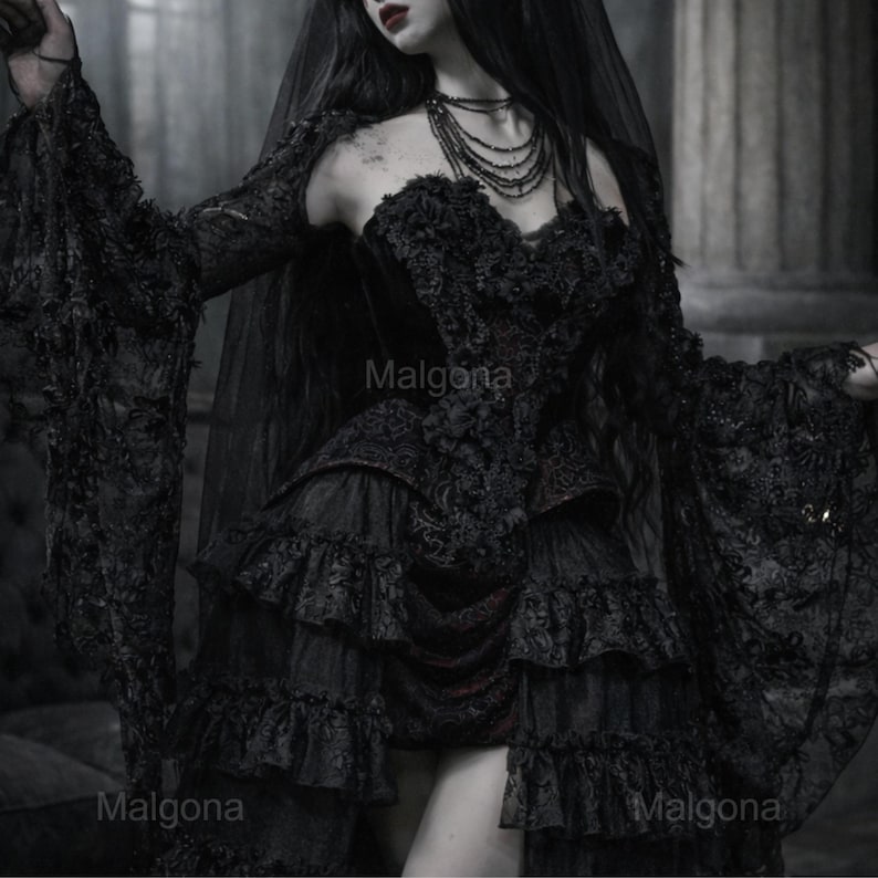 May include: A black dress featuring long flared lace sleeves, a sweetheart neckline, and a multi-layered ruffled skirt. The bodice is adorned with intricate lace detailing and floral textures. A sheer black veil is draped over the shoulders.