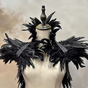 May include: A dramatic black feather shoulder piece displayed on a mannequin. The piece features layers of dark feathers and a structured design, with straps across the shoulders. The word "Malgona" is visible in the upper right corner.