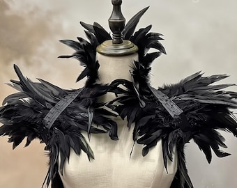 May include: A dramatic black feather shoulder piece displayed on a mannequin. The piece features layers of dark feathers and a structured design, with straps across the shoulders. The word "Malgona" is visible in the upper right corner.