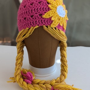 Puede incluir: Un gorro de ganchillo hecho a mano en color rosa vibrante, adornado con un girasol amarillo y largas extensiones de cabello trenzado de hilo amarillo y marrón. El gorro está diseñado para parecerse al peinado de un personaje.
