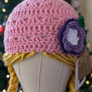 Pode incluir: Chapéu de crochê rosa com uma flor roxa e folhas verdes. O chapéu tem duas tranças longas amarelas presas na parte inferior.