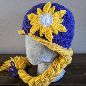 Pode incluir: Chapéu de crochê roxo e amarelo com uma trança amarela presa. O chapéu apresenta um girassol amarelo com um centro branco.