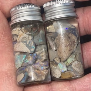 Könnte beinhalten: Zwei kleine Glasflaschen mit silbernen Deckeln, gefüllt mit einer Vielzahl farbenfroher, schillernder Opalfragmente. Die Opale zeigen Farbtöne von Blau, Grün und Lila, gemischt mit anderen Steinfragmenten.