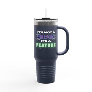May include: Navy blue travel mug with a handle and a straw. The mug features the text "IT'S NOT A BUG IT'S A FEATURE" in a pixelated font. The mug has a silver rim and a black lid.